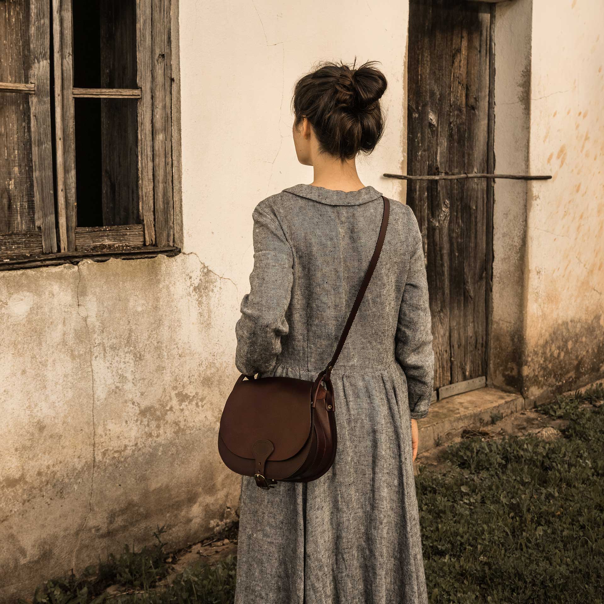 Une femme porte en bandoulière une besace en cuir devant une vieille maison