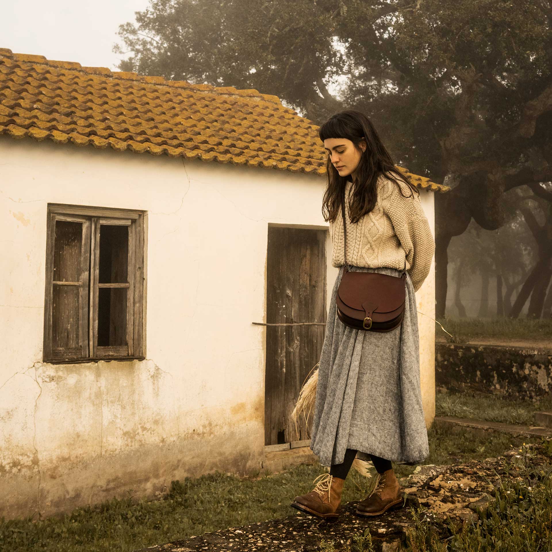 Une femme porte en bandoulière une besace en cuir devant une vieille maison