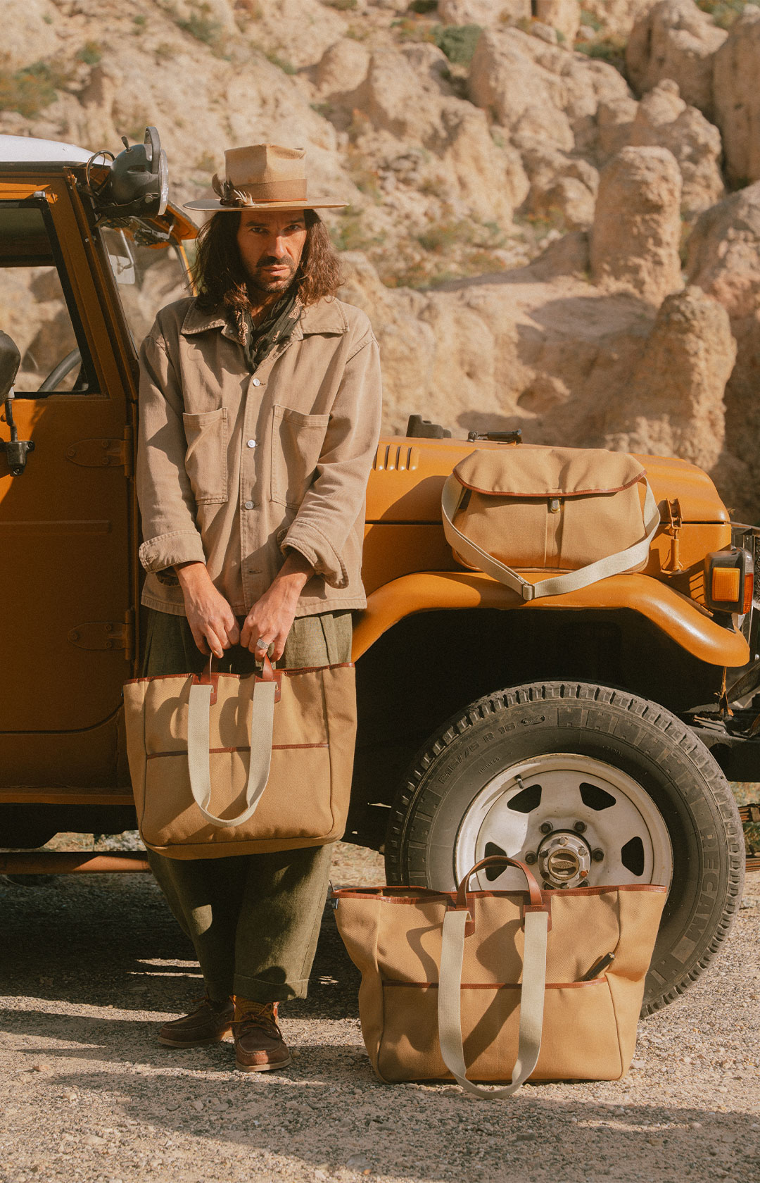한 남자가 지프차 앞에 서서 캔버스 가방을 들고 있다. 그 옆에는 다른 캔버스 가방 두 개가 놓여 있다.