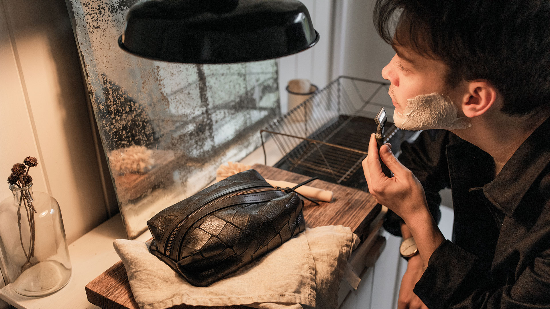 A man shaves in front of a mirror, and on the sink is a leather toiletry bag