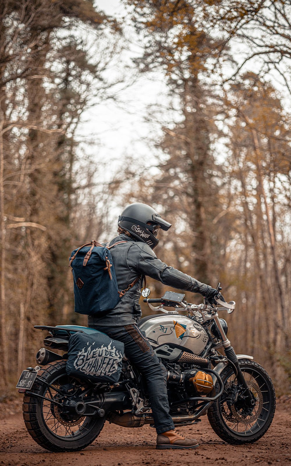 A man on a motorcycle with a canvas rucksack and a motorcycle bag attached to the back.