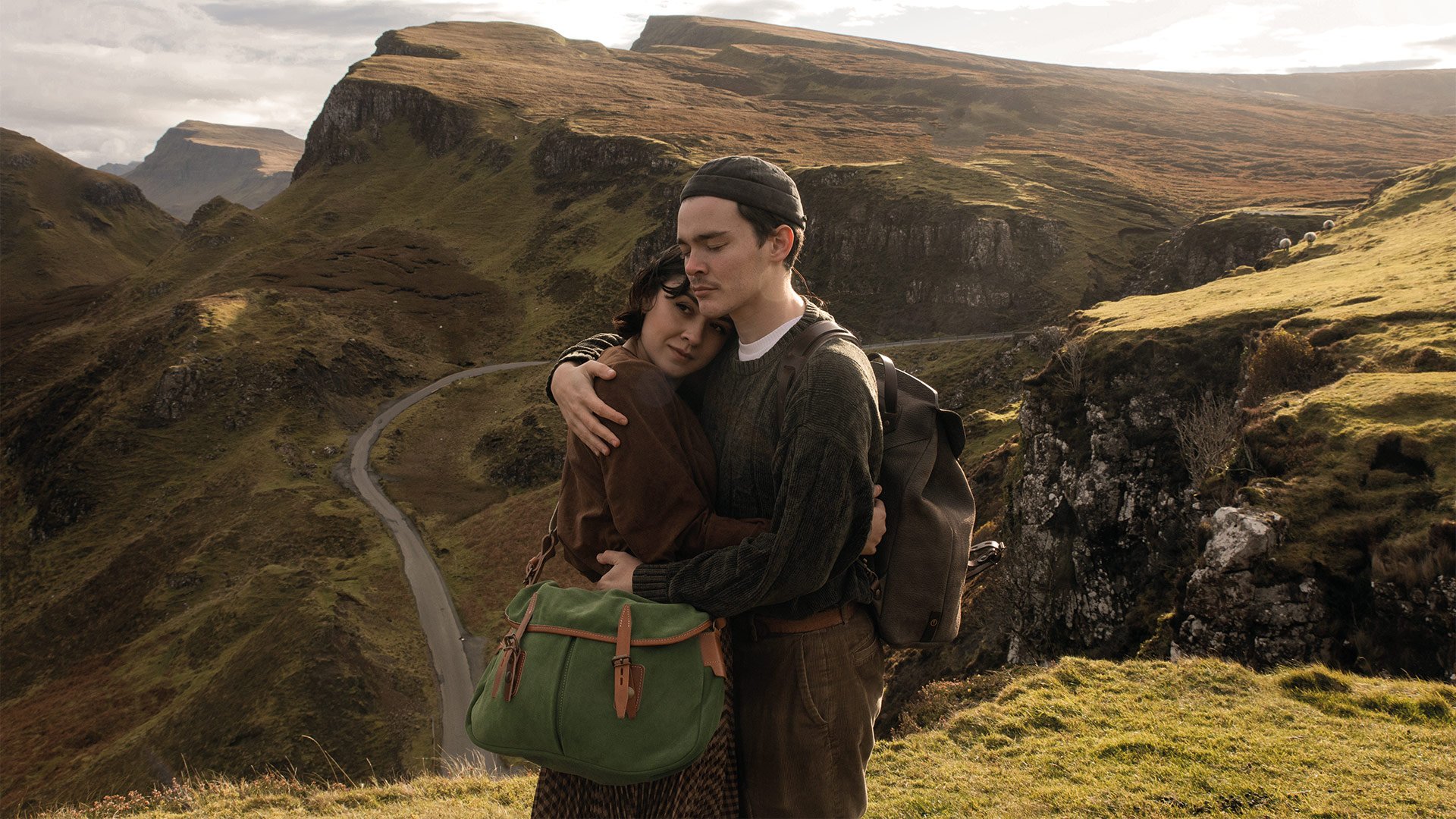 In a mountainous landscape, a man is carrying a leather backpack and a woman is carrying a suede pouch. The two give each other a hug.