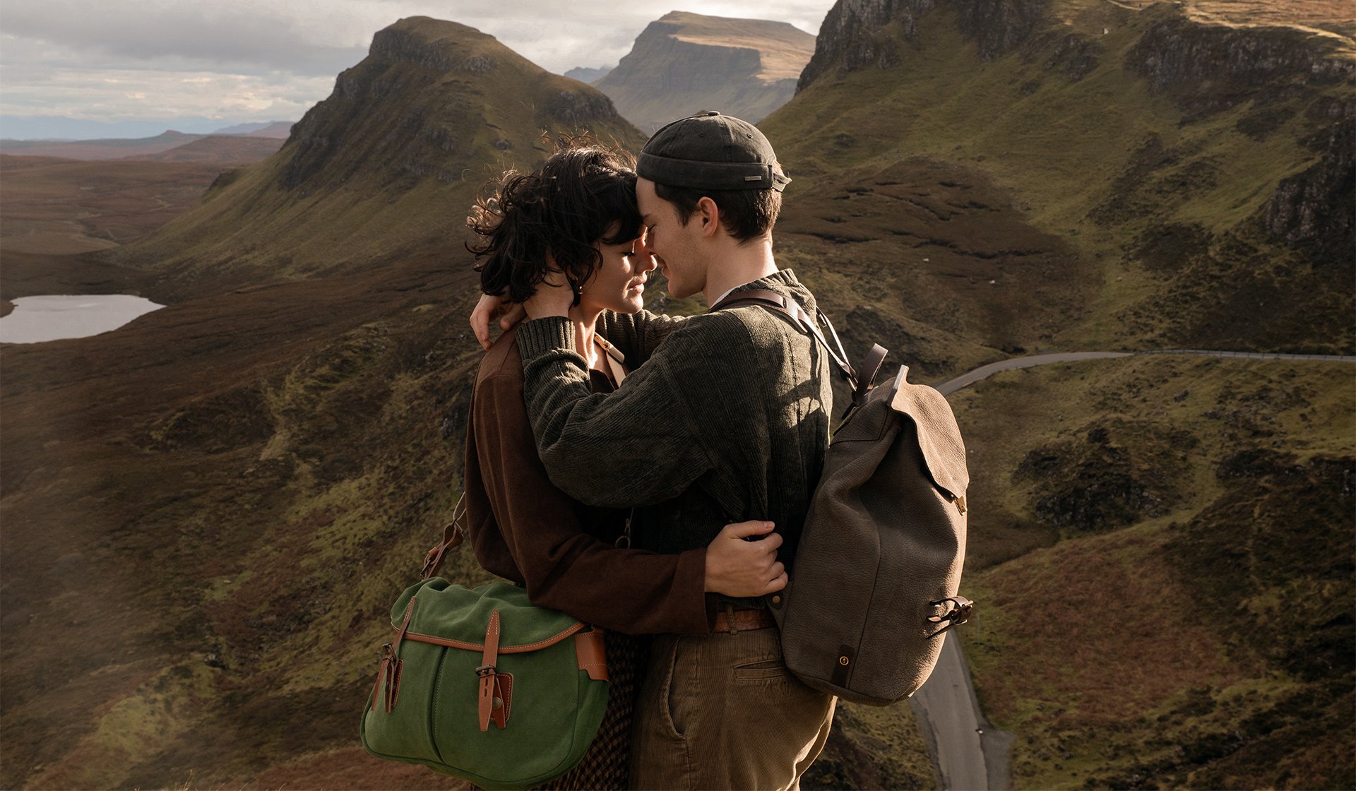 Dans un paysage montagneux, un homme porte un sac a dos en cuir et une femme porte une besace en cuir velours. Les deux se font un calins 
