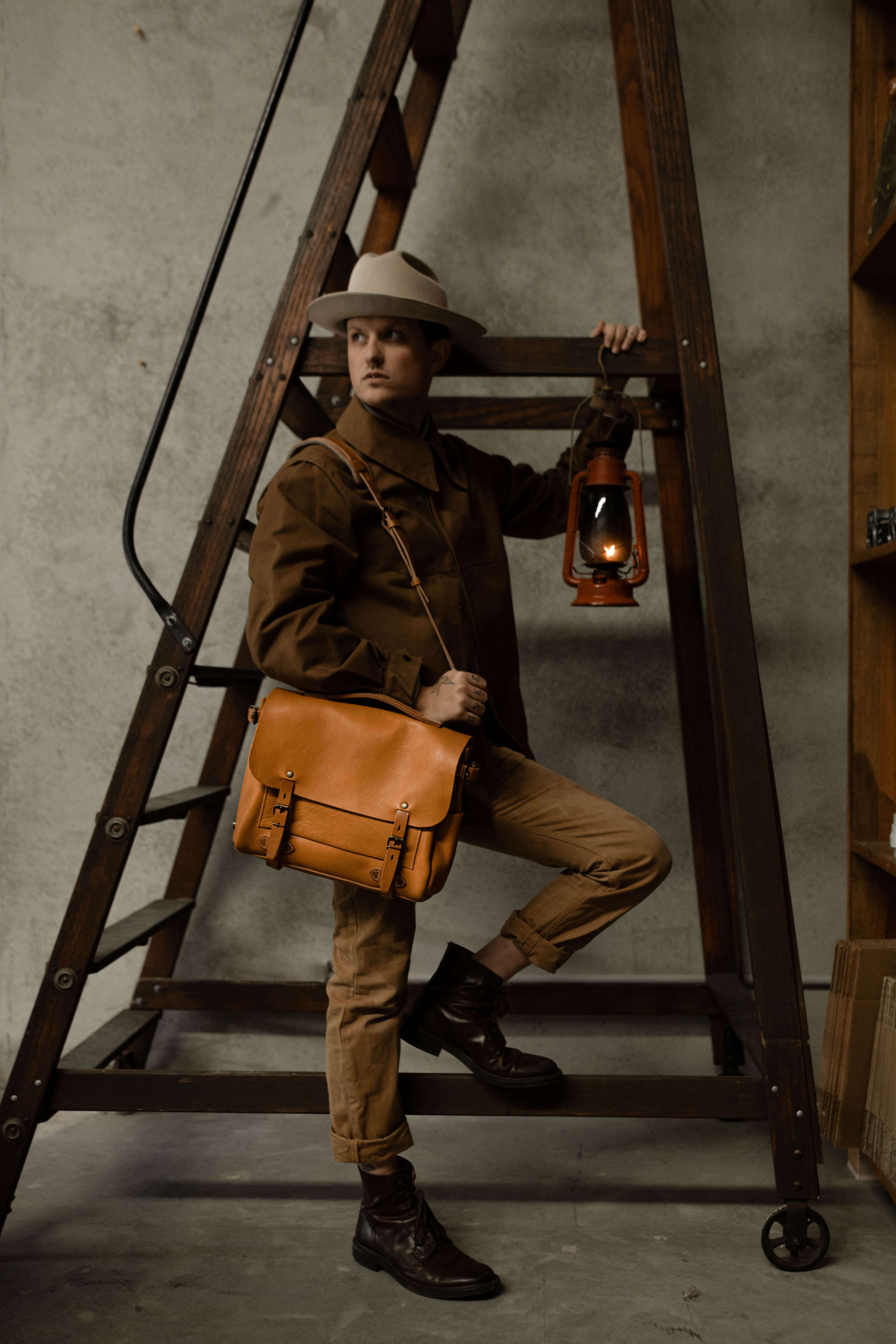 A man slings a leather shoulder bag over his shoulder in front of a wooden stepladder.