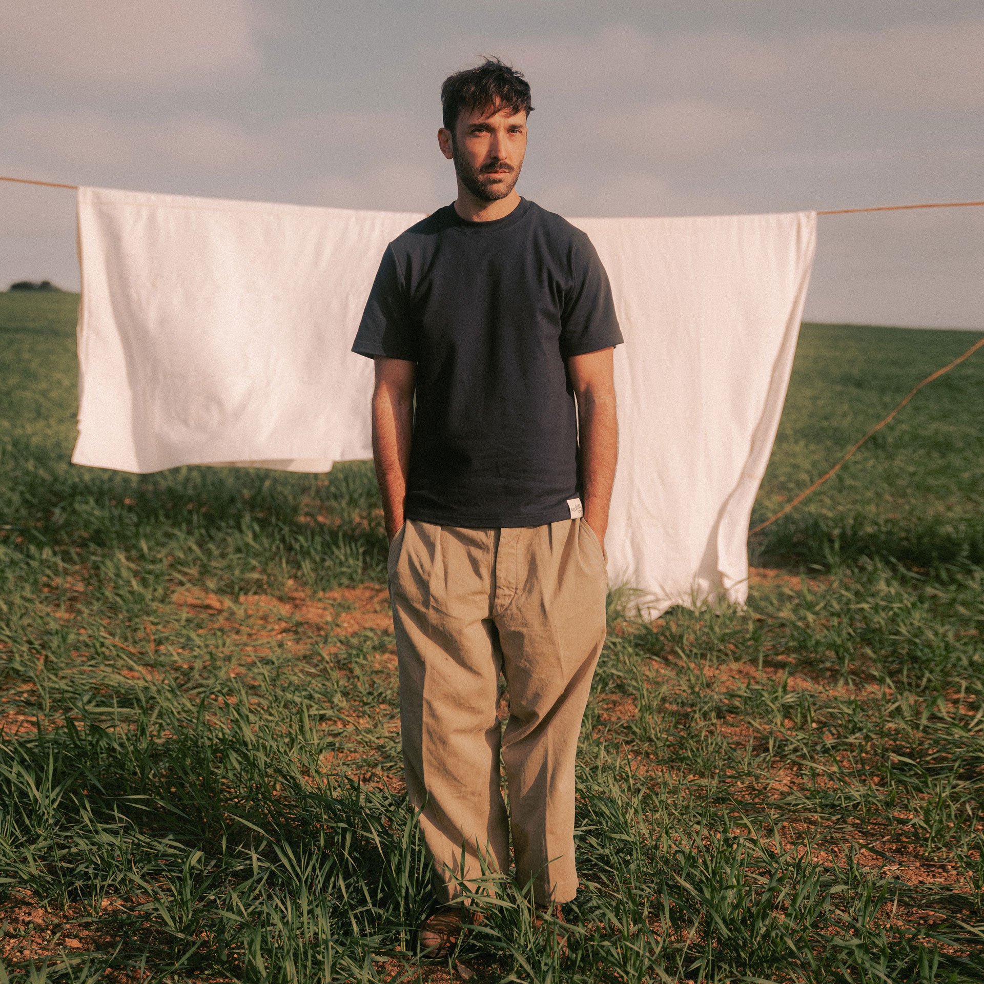 Un homme porte un t-shirt en coton bio bleu devant un fil de linge avec draps blanc