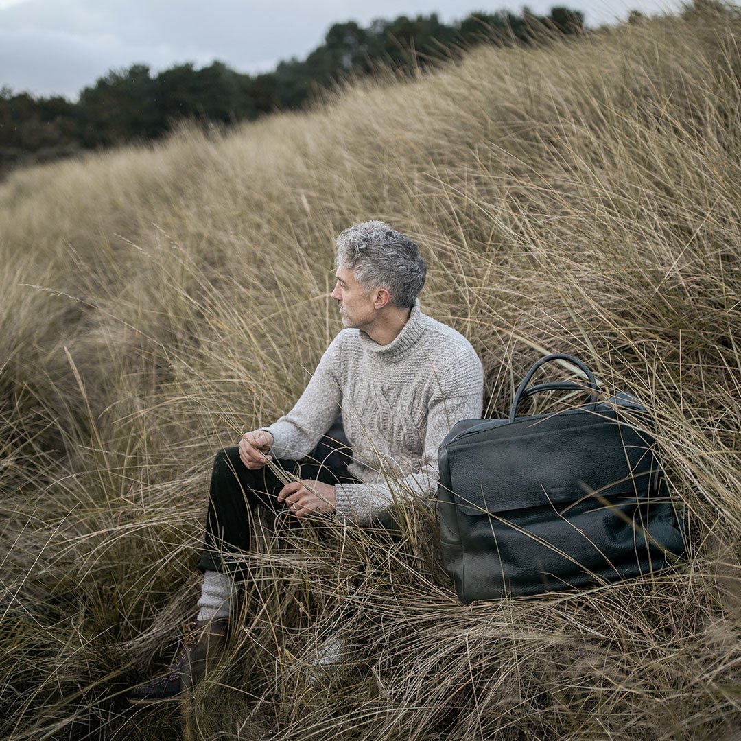 Un homme est assis dans des hautes herbes et pose avec un sac en cuir