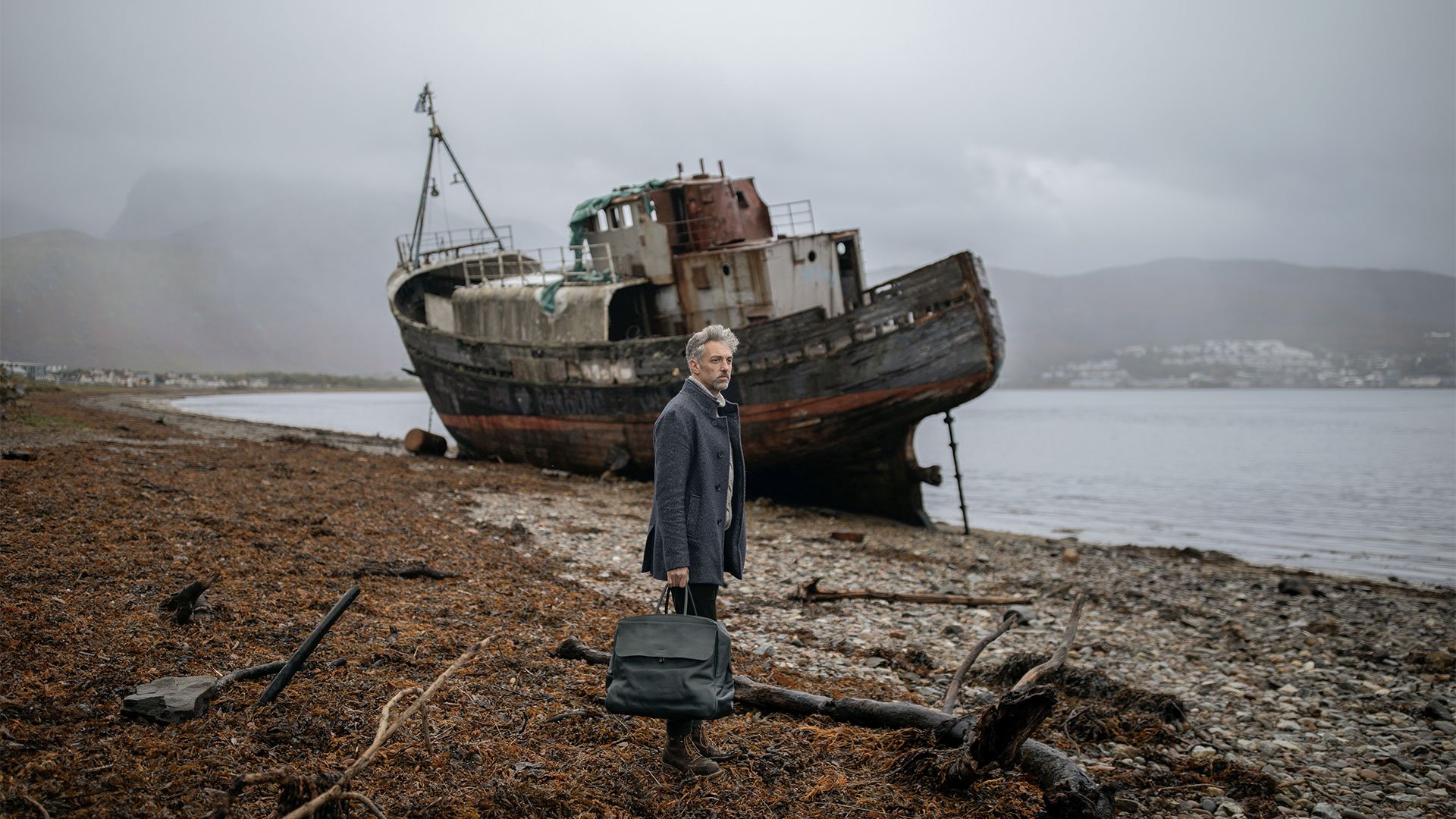 A man carries a leather bag in front of a shipwreck