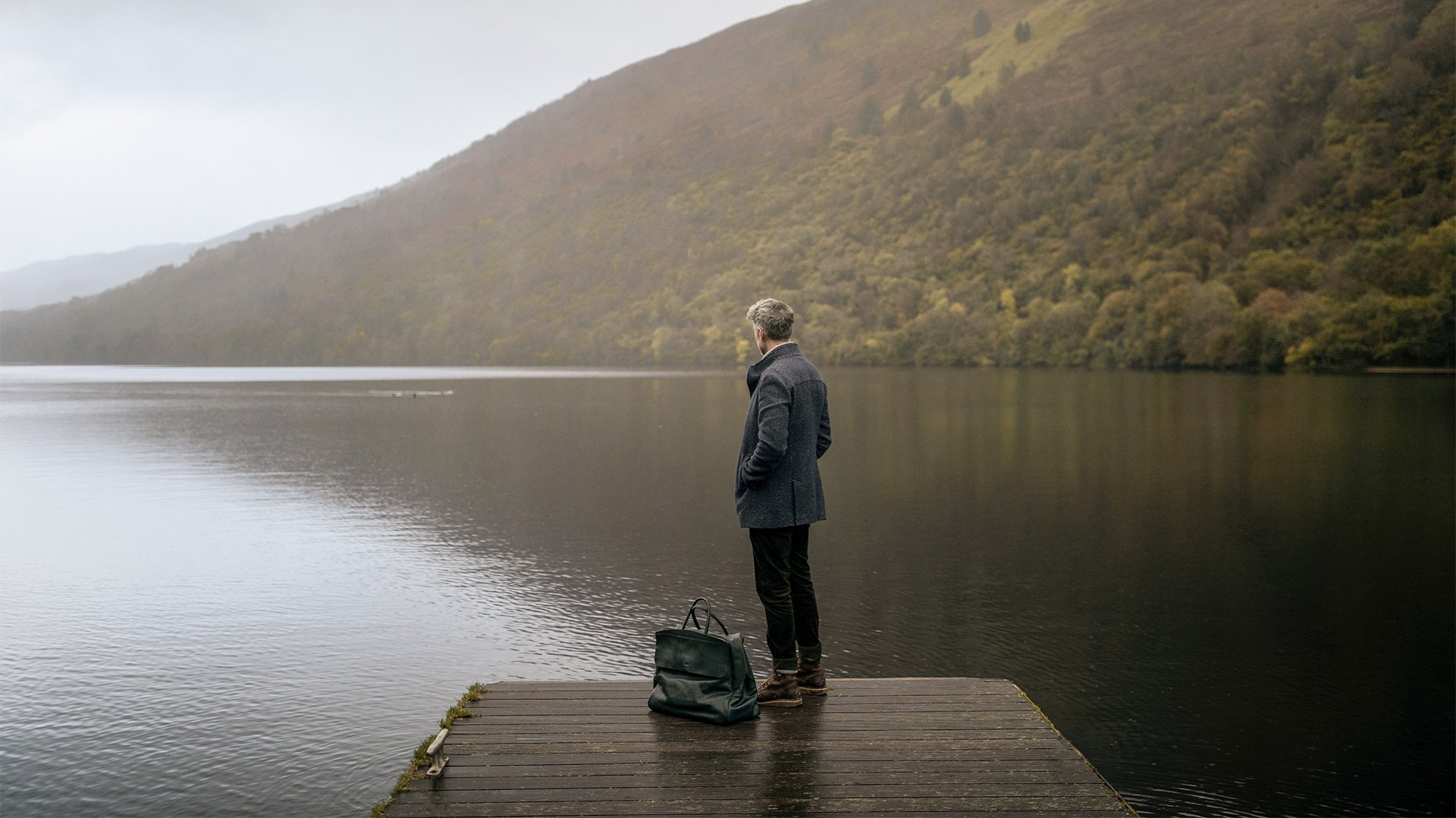 A man on a pontoon next to his travel bag looks out over a lake