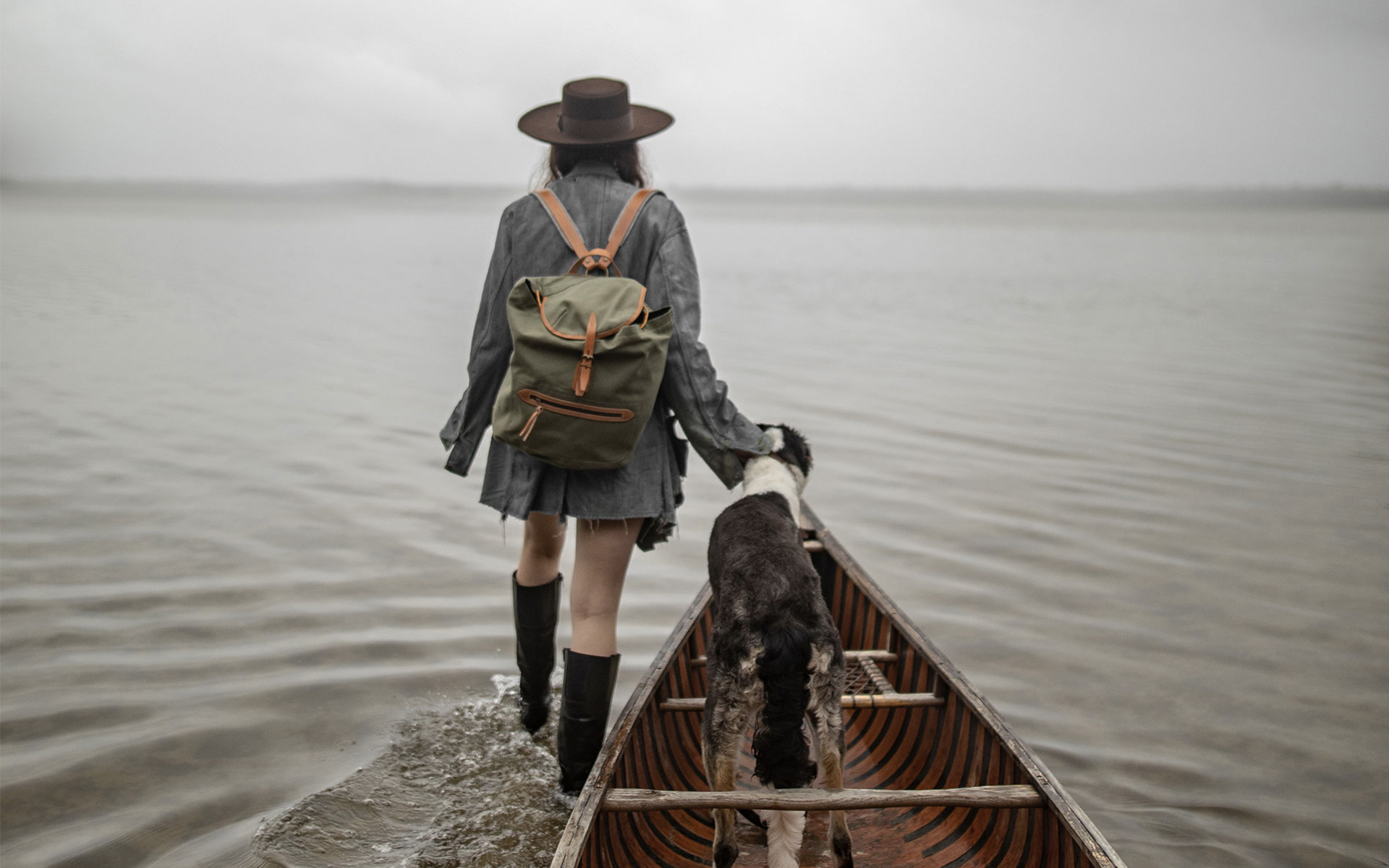 A woman walks through the water, carrying a canvas backpack and towing a boat